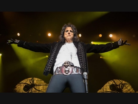 Alice Cooper performing on stage in his black stage costume with skull belt buckle, arms outstretched under green lighting