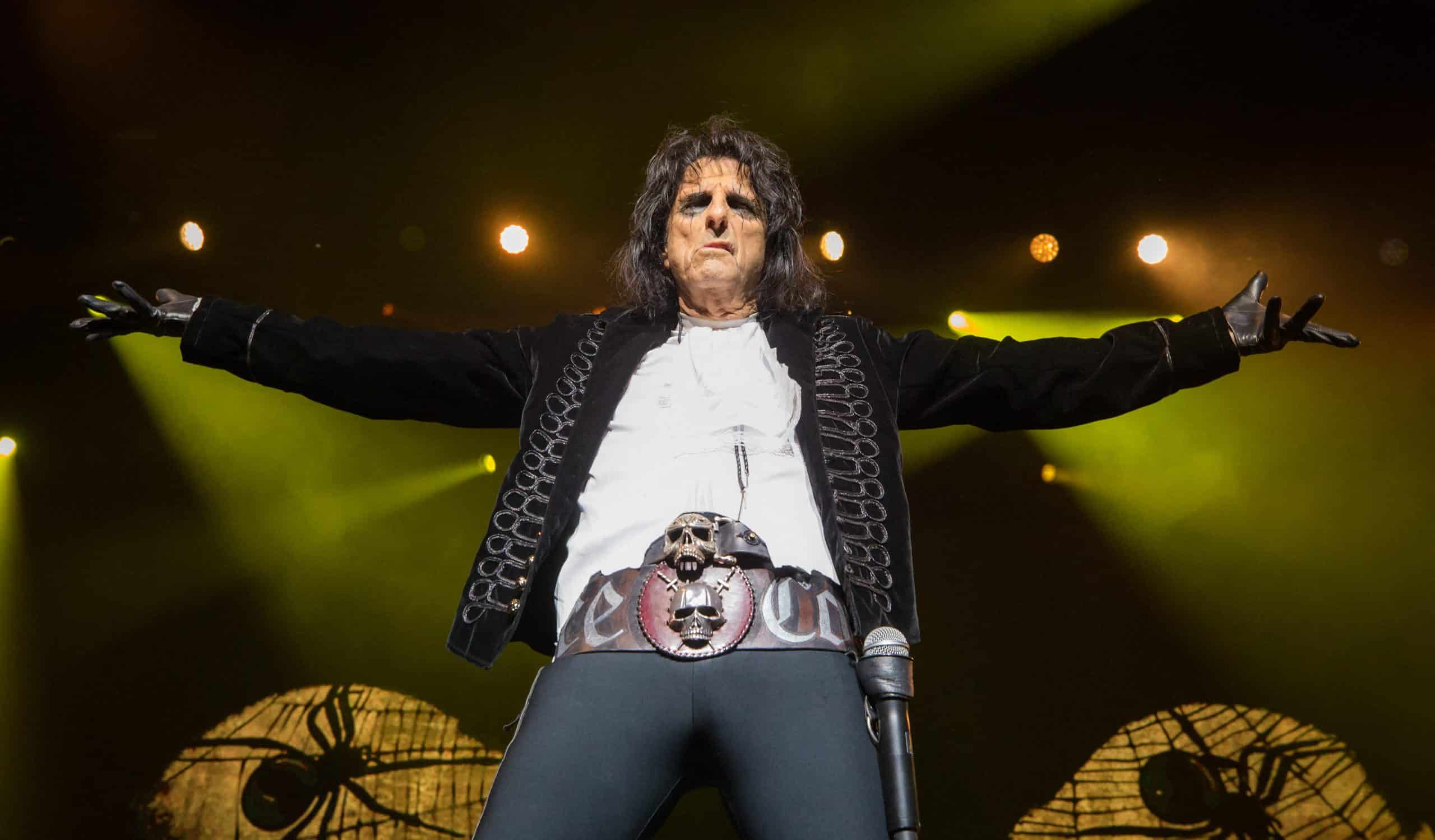 Alice Cooper performing on stage in his black stage costume with skull belt buckle, arms outstretched under green lighting