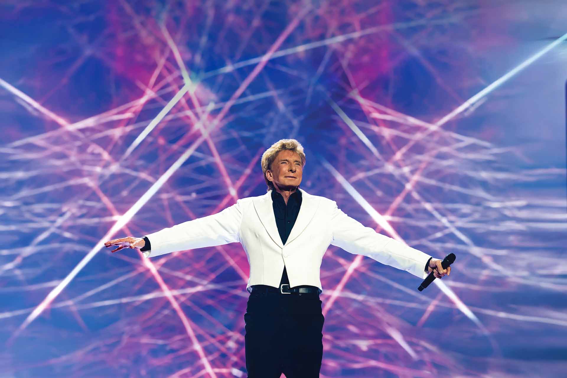 Barry Manilow in a white jacket holding a microphone on stage with pink and blue light beams behind him
