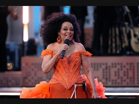 Diana Ross sings into a handheld microphone on stage wearing an orange beaded gown with ruffled sleeves