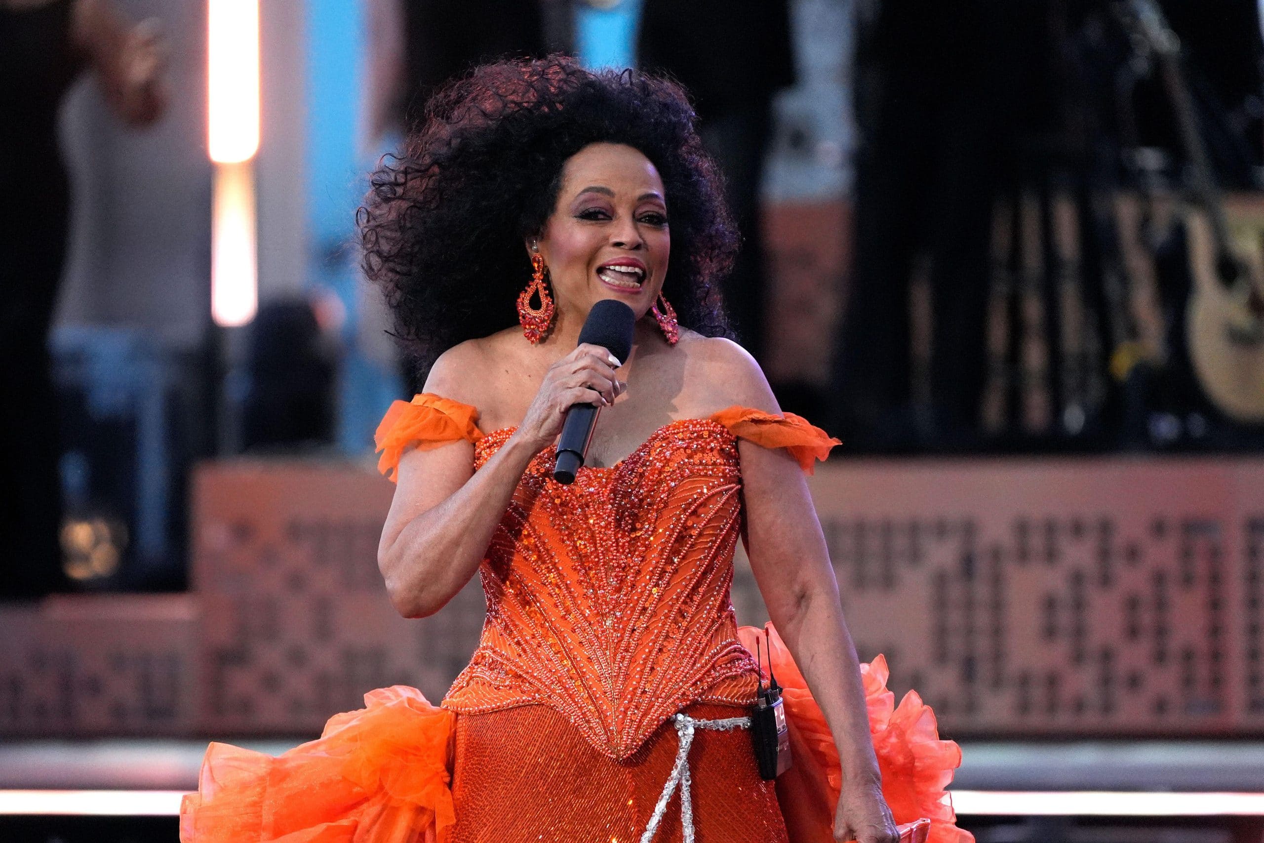 Diana Ross sings into a handheld microphone on stage wearing an orange beaded gown with ruffled sleeves