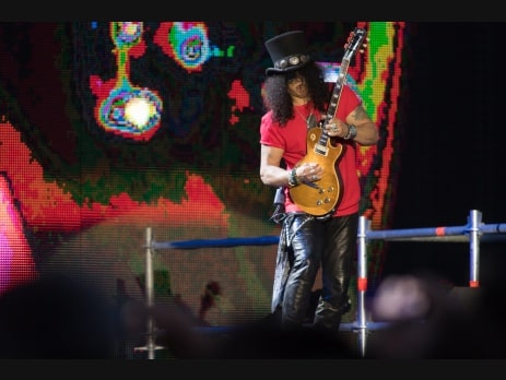 Slash of Guns N' Roses plays a Gibson Les Paul on stage in a black top hat and red shirt against a colorful LED backdrop