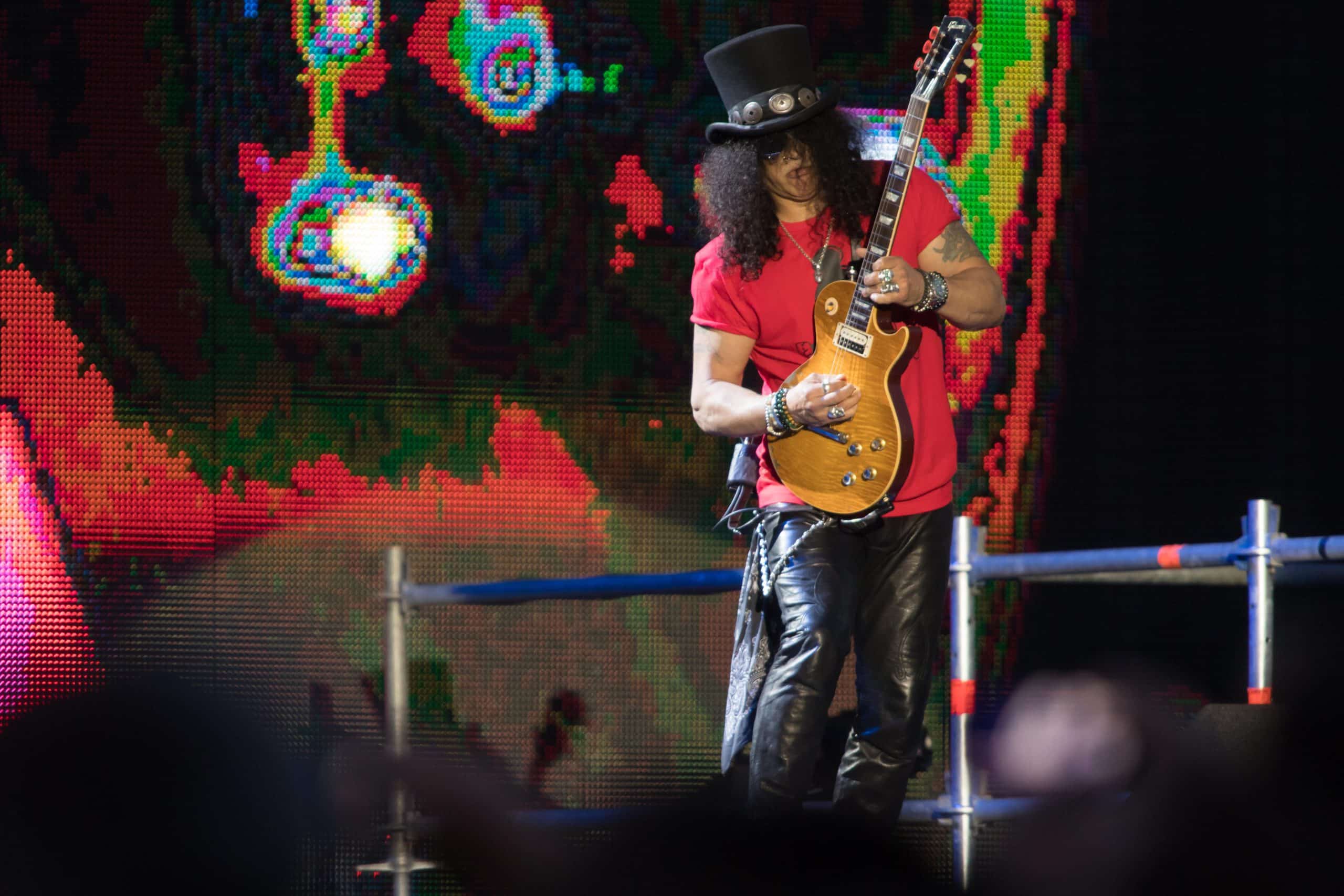 Slash of Guns N' Roses plays a Gibson Les Paul on stage in a black top hat and red shirt against a colorful LED backdrop