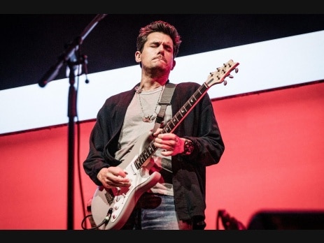John Mayer playing a white electric guitar on stage against a red and white backdrop