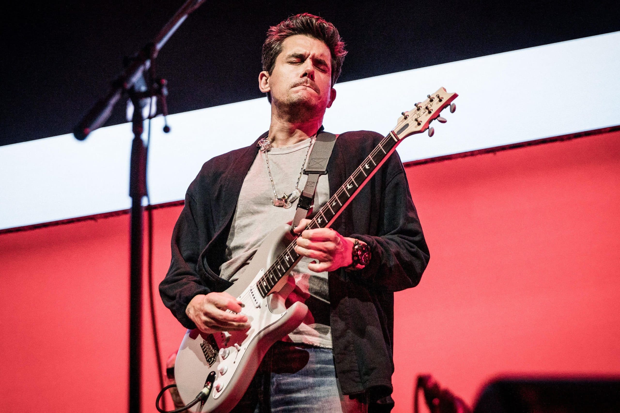 John Mayer playing a white electric guitar on stage against a red and white backdrop