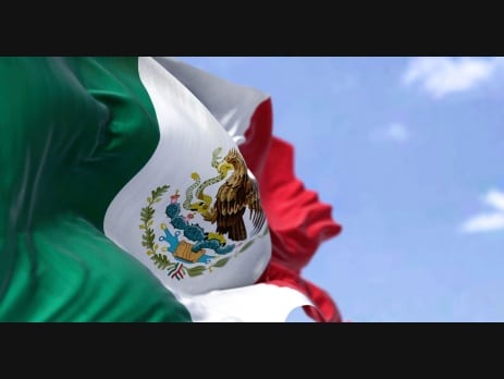 The flag of Mexico waving in the wind against a blue sky, with the national coat of arms of an eagle and serpent at its center