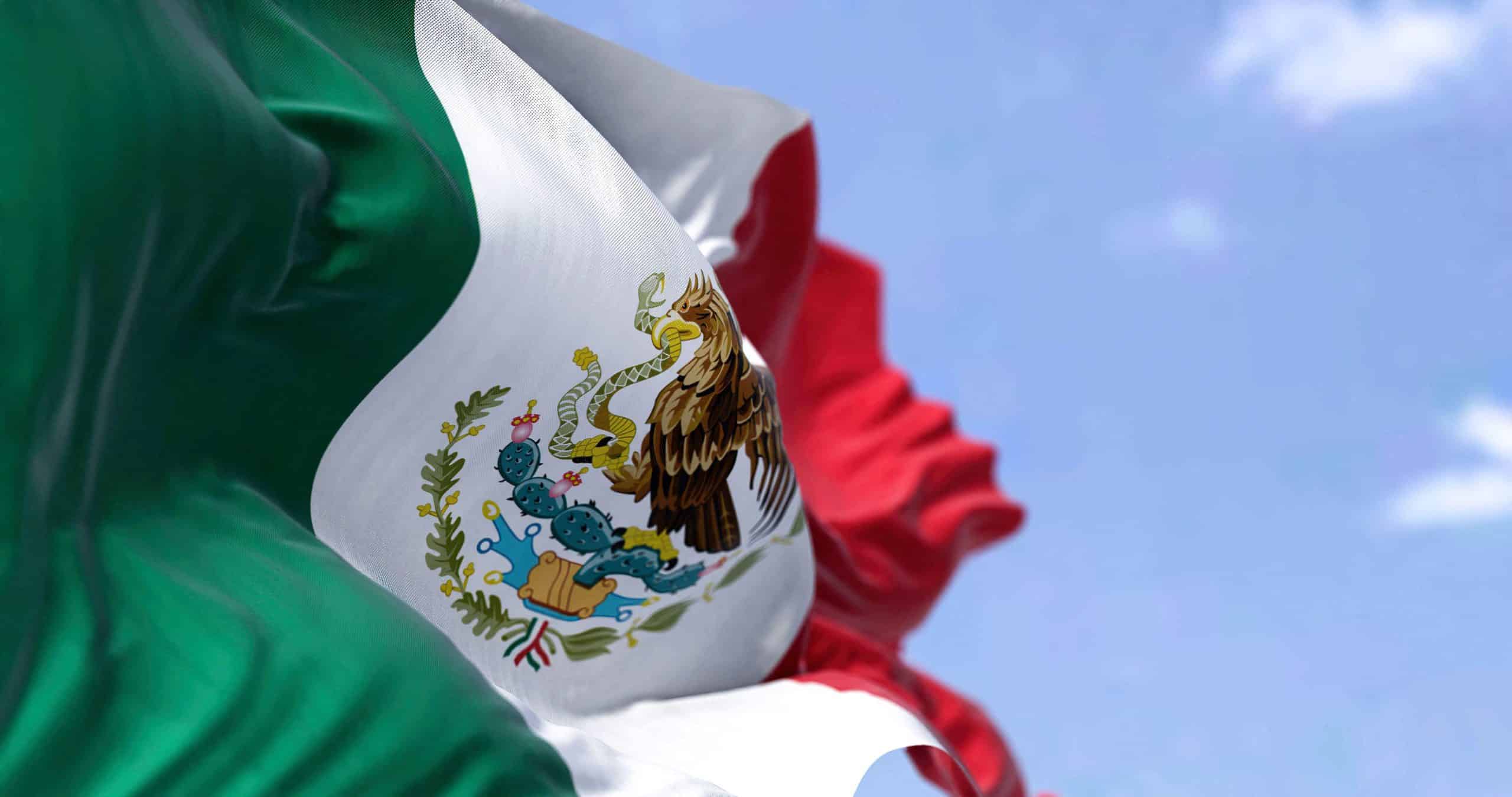 The flag of Mexico waving in the wind against a blue sky, with the national coat of arms of an eagle and serpent at its center