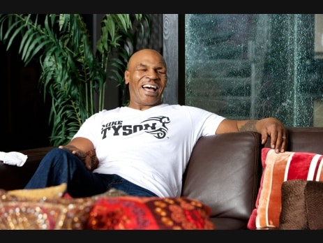 Mike Tyson laughing while seated indoors wearing a Mike Tyson branded t-shirt