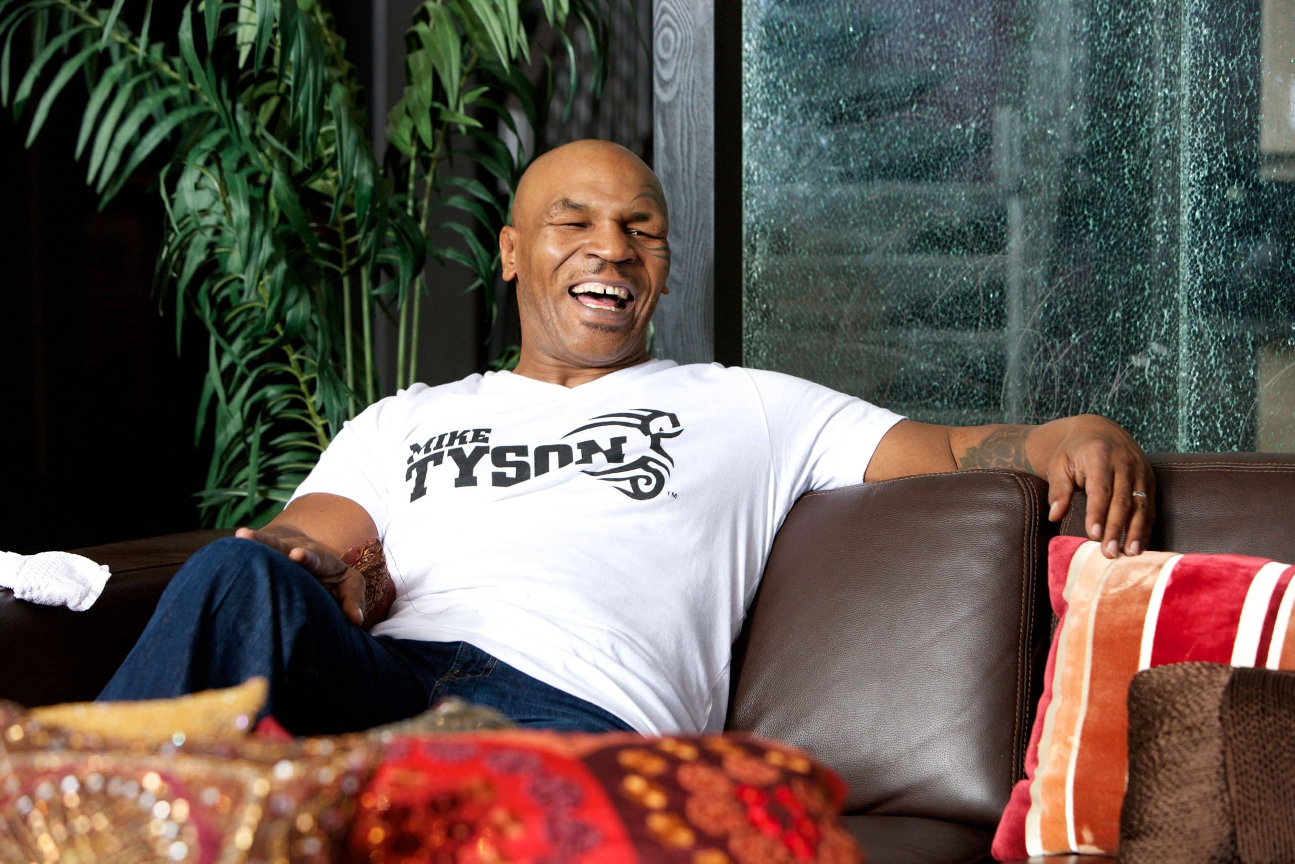 Mike Tyson laughing while seated indoors wearing a Mike Tyson branded t-shirt