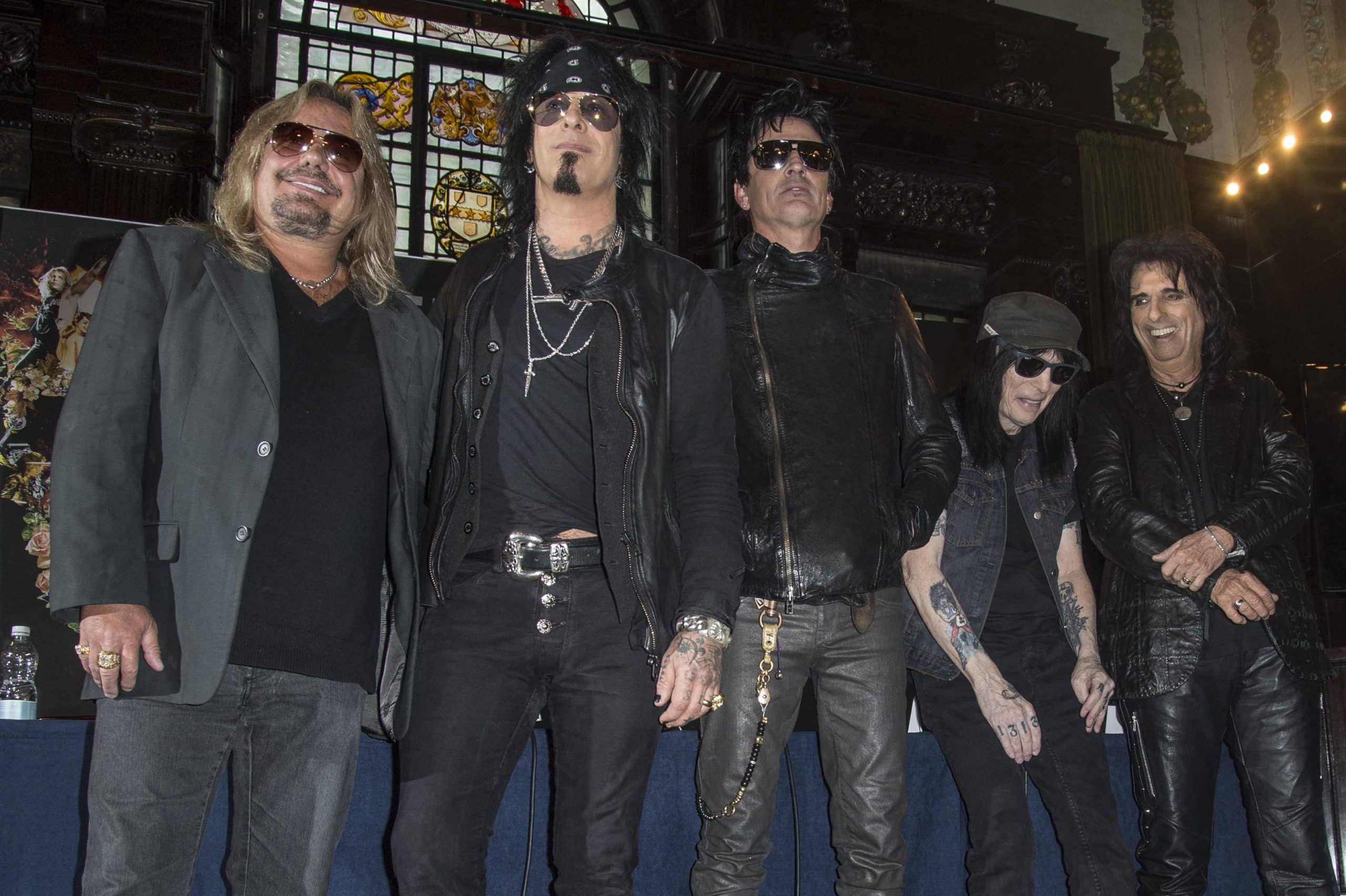 The five members of Motley Crue standing together in a row in sunglasses and black leather in front of a stained-glass window