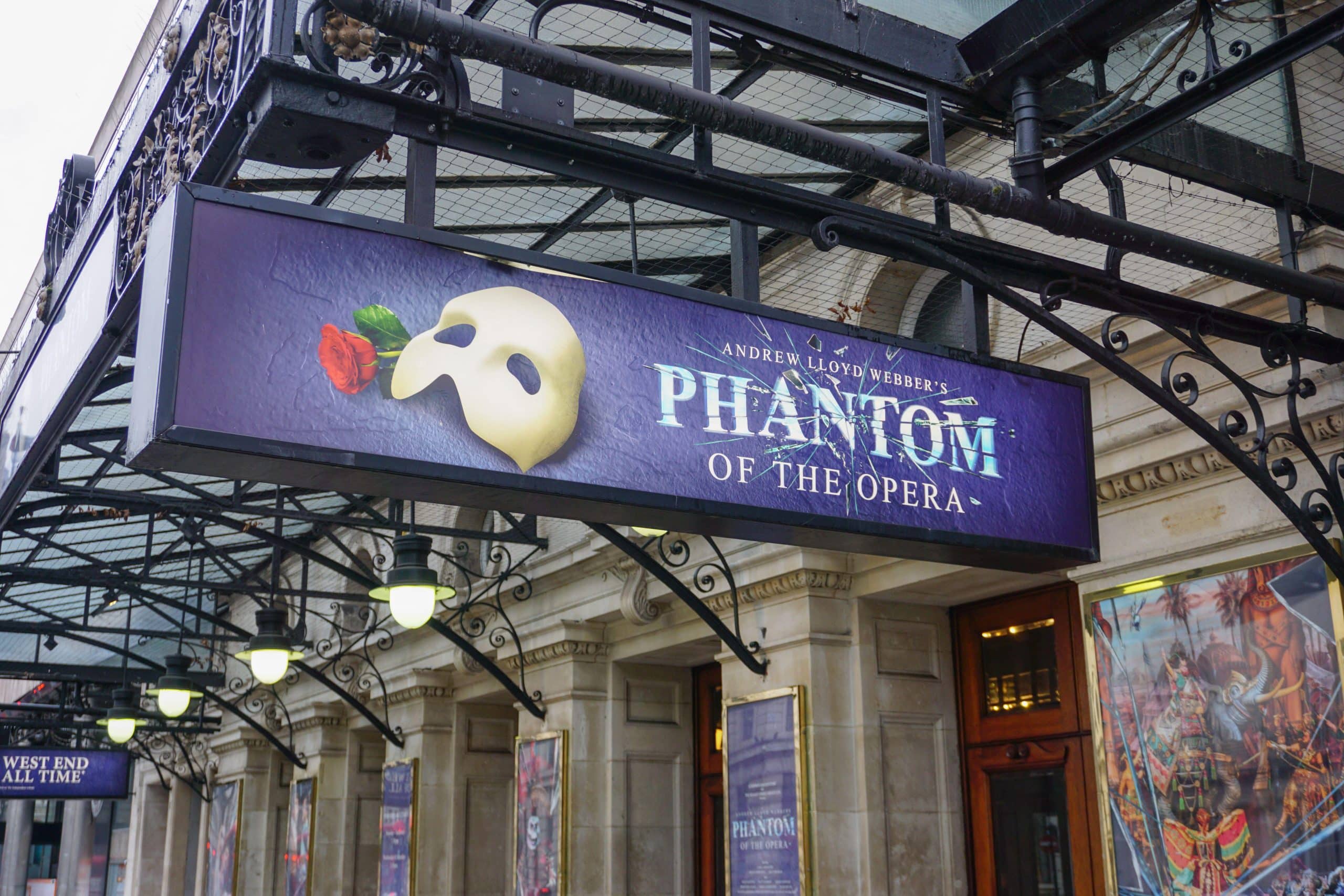 Purple theatre marquee sign reading Andrew Lloyd Webber's Phantom of the Opera, beside a white half mask and a red rose