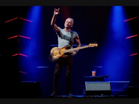 Sting performing on stage with a bass guitar, right arm raised, under blue stage lights