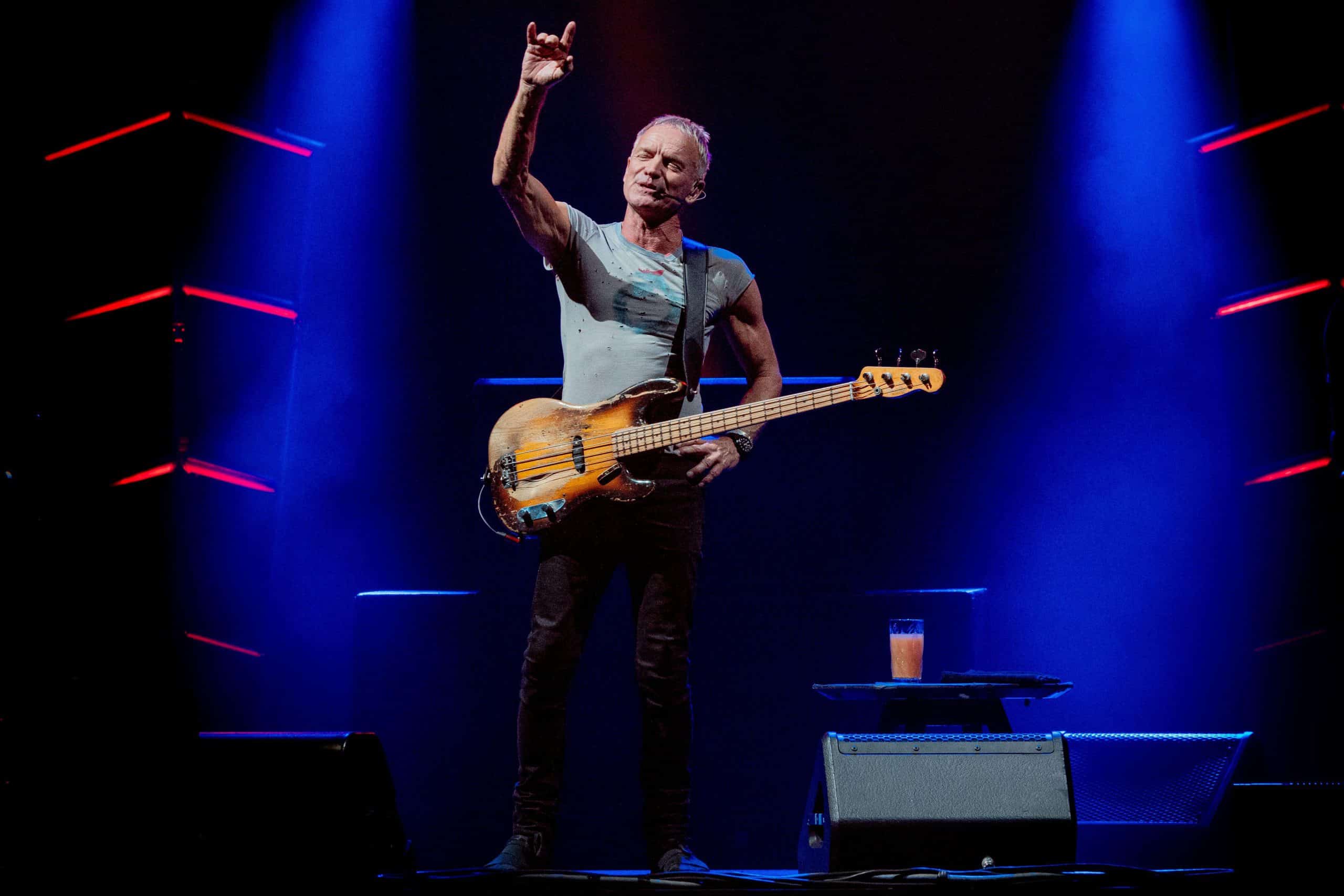 Sting performing on stage with a bass guitar, right arm raised, under blue stage lights