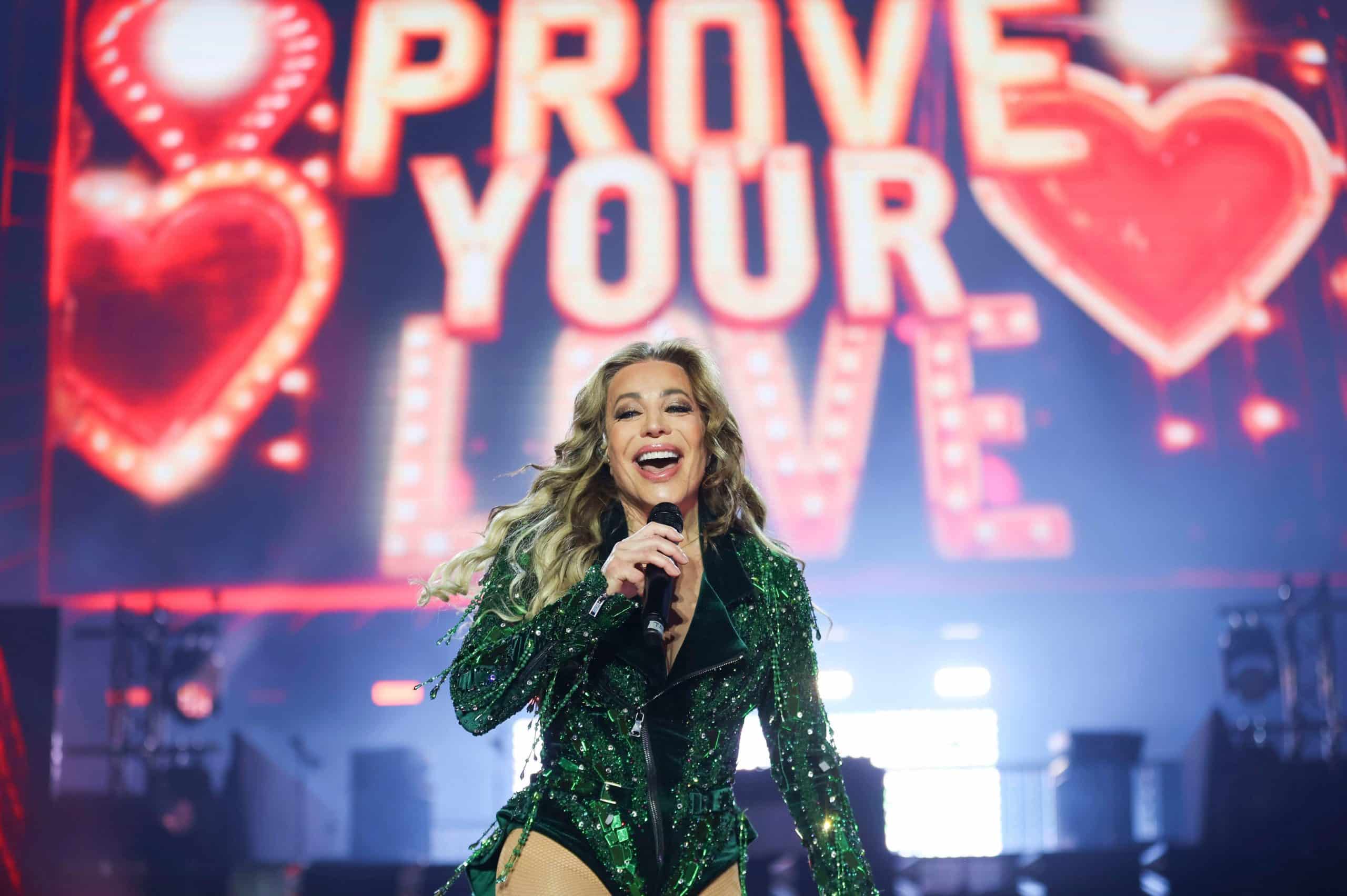Taylor Dayne performing in a green sequined outfit in front of a Prove Your Love stage backdrop