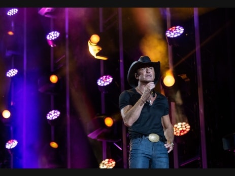Tim McGraw performing on stage in a black cowboy hat and black shirt under purple and gold concert lighting