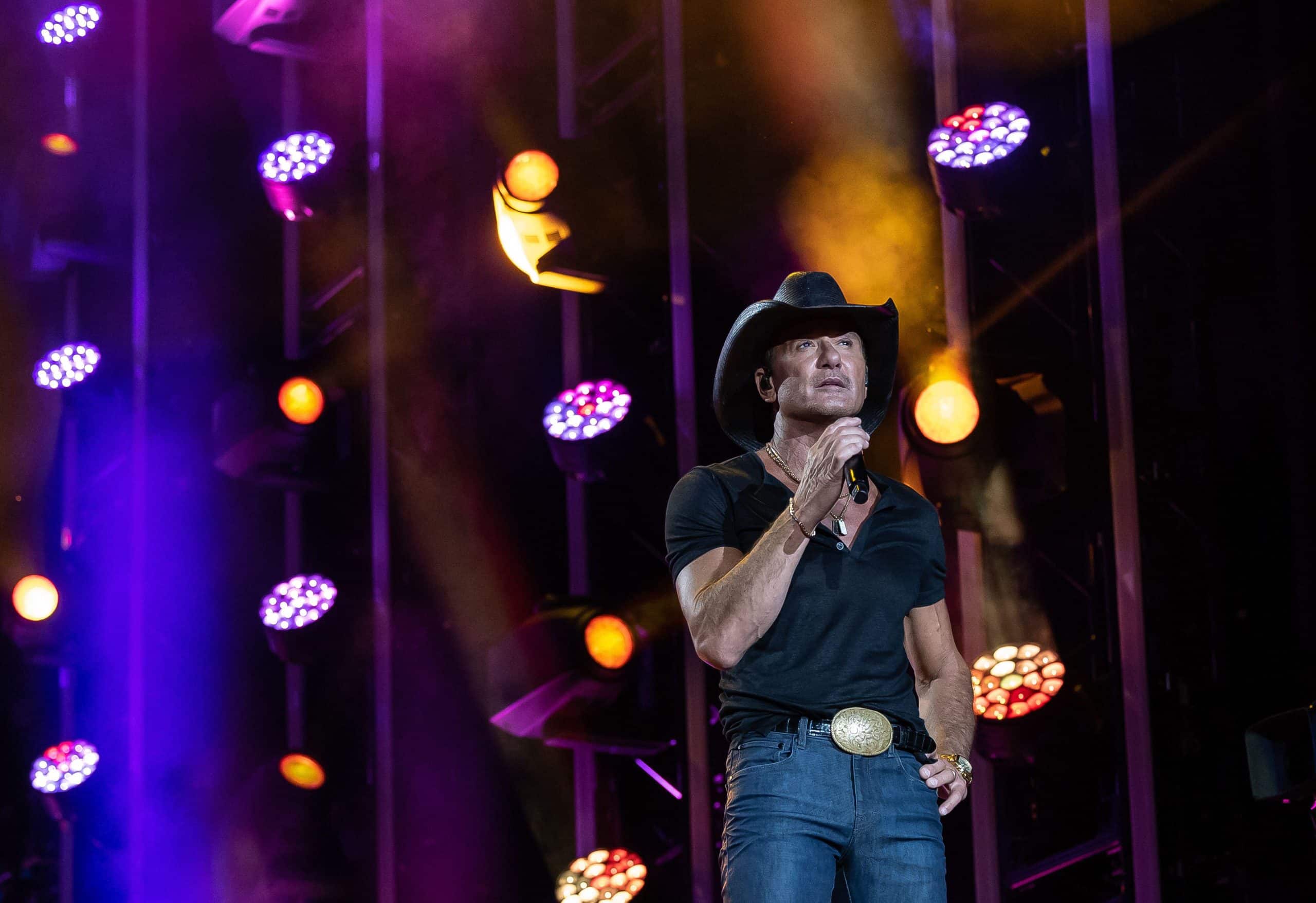 Tim McGraw performing on stage in a black cowboy hat and black shirt under purple and gold concert lighting