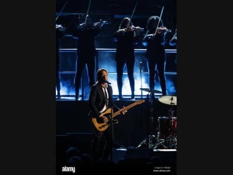 Keith Urban plays guitar on stage at a concert, with string musicians performing behind him under blue stage lights