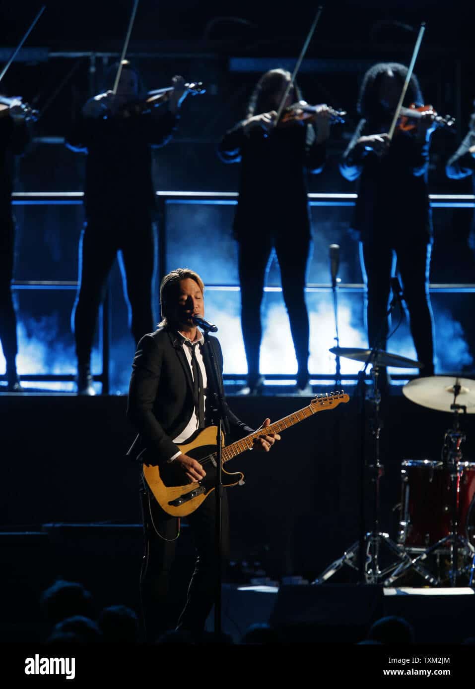 Keith Urban plays guitar on stage at a concert, with string musicians performing behind him under blue stage lights