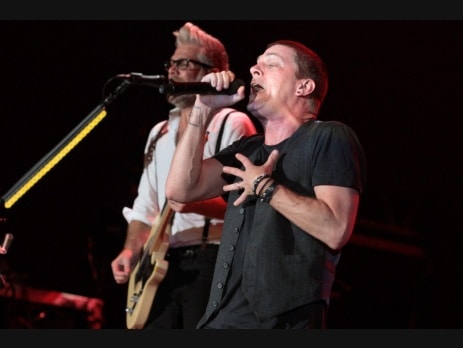 Matchbox Twenty performing live on a dark stage, the singer at a microphone in the foreground with a guitarist behind him