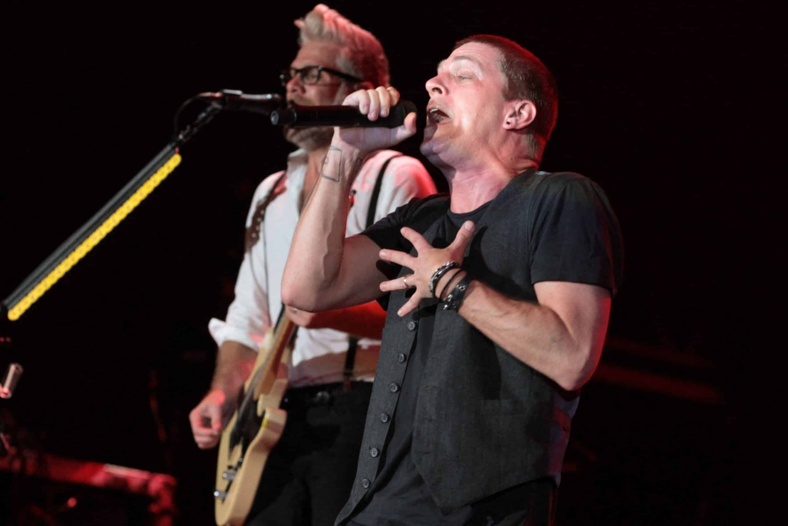 Matchbox Twenty performing live on a dark stage, the singer at a microphone in the foreground with a guitarist behind him