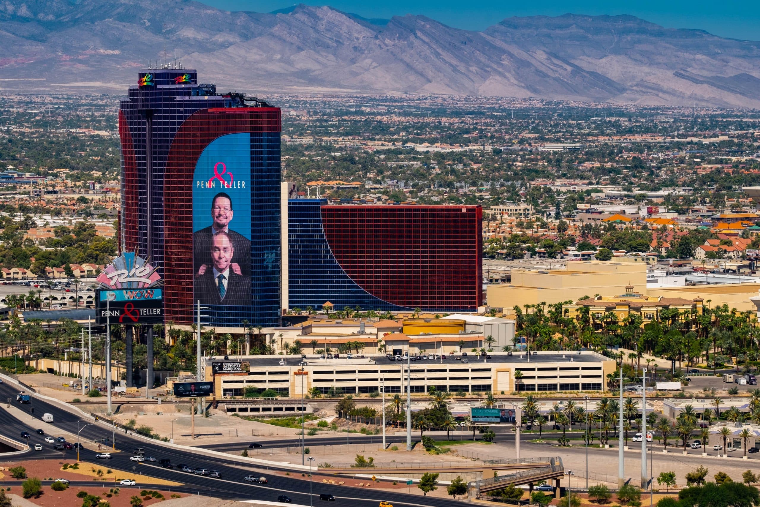 The Rio All-Suite Hotel tower in Las Vegas wrapped with a large Penn and Teller advertisement, with the resort marquee and desert mountains in the background