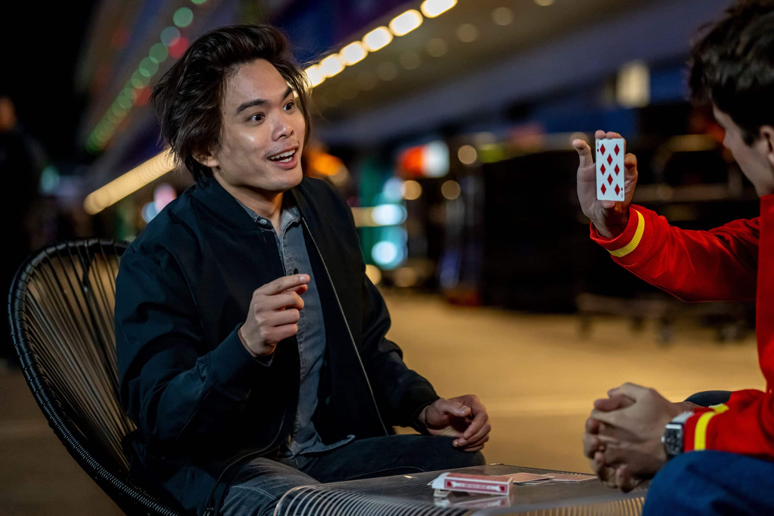 Shin Lim performs close-up card magic for a spectator outdoors at night, with a playing card held up between them